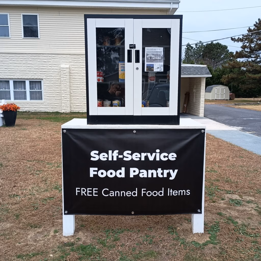 Outdoor self-service food pantry cabinet with glass doors stocked with canned goods and a bold Self-Service Food Pantry sign below.